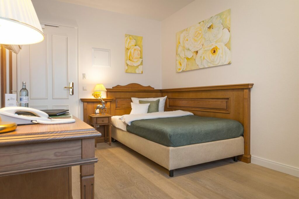 Gemütlich eingerichtetes Einzelzimmer im Landhaus Lellichow mit Holzmöbeln, einem Bett mit grüner Tagesdecke und Bildern von Rosen an der Wand.