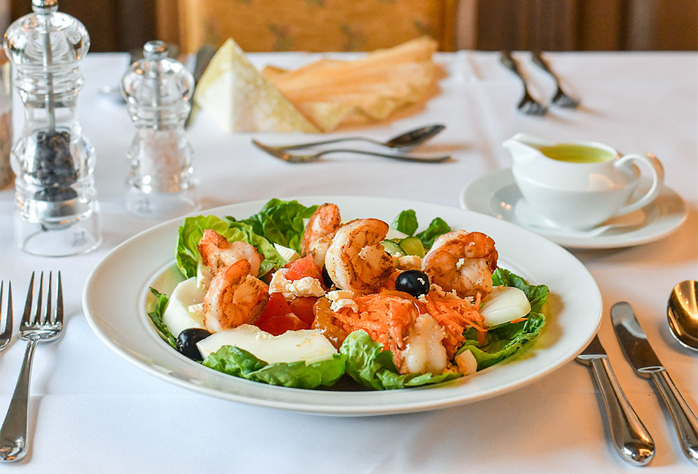 Ein frisch angerichteter Salatteller mit gebratenen Garnelen, Feta und Gemüse auf einem weiß gedeckten Restauranttisch.