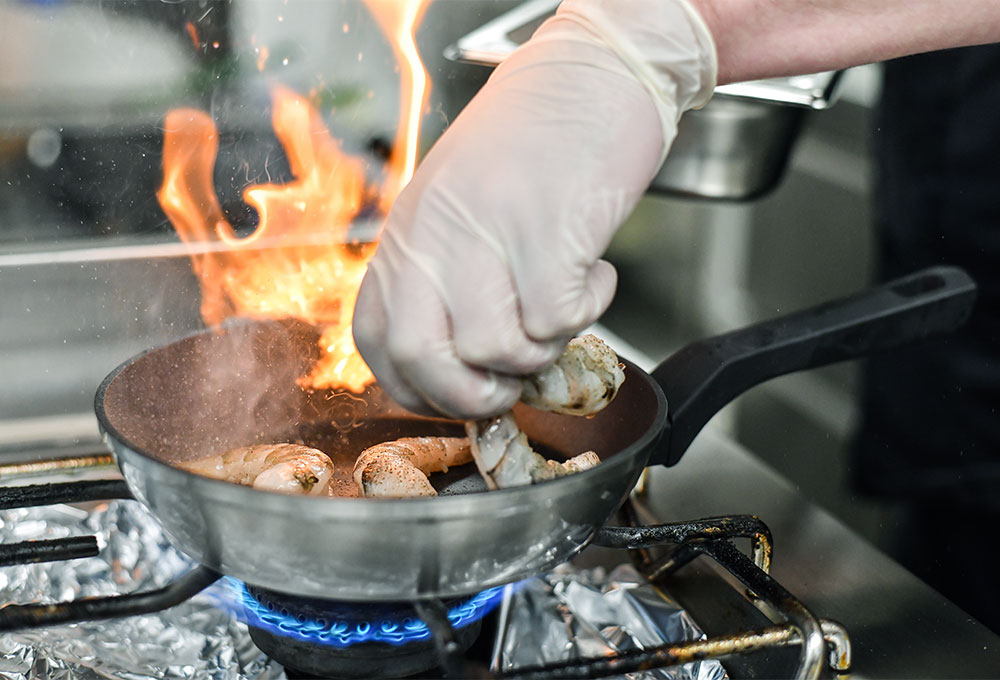 Ein Koch legt mit einer behandschuhten Hand Garnelen in eine heiße Pfanne, aus der Flammen schlagen.