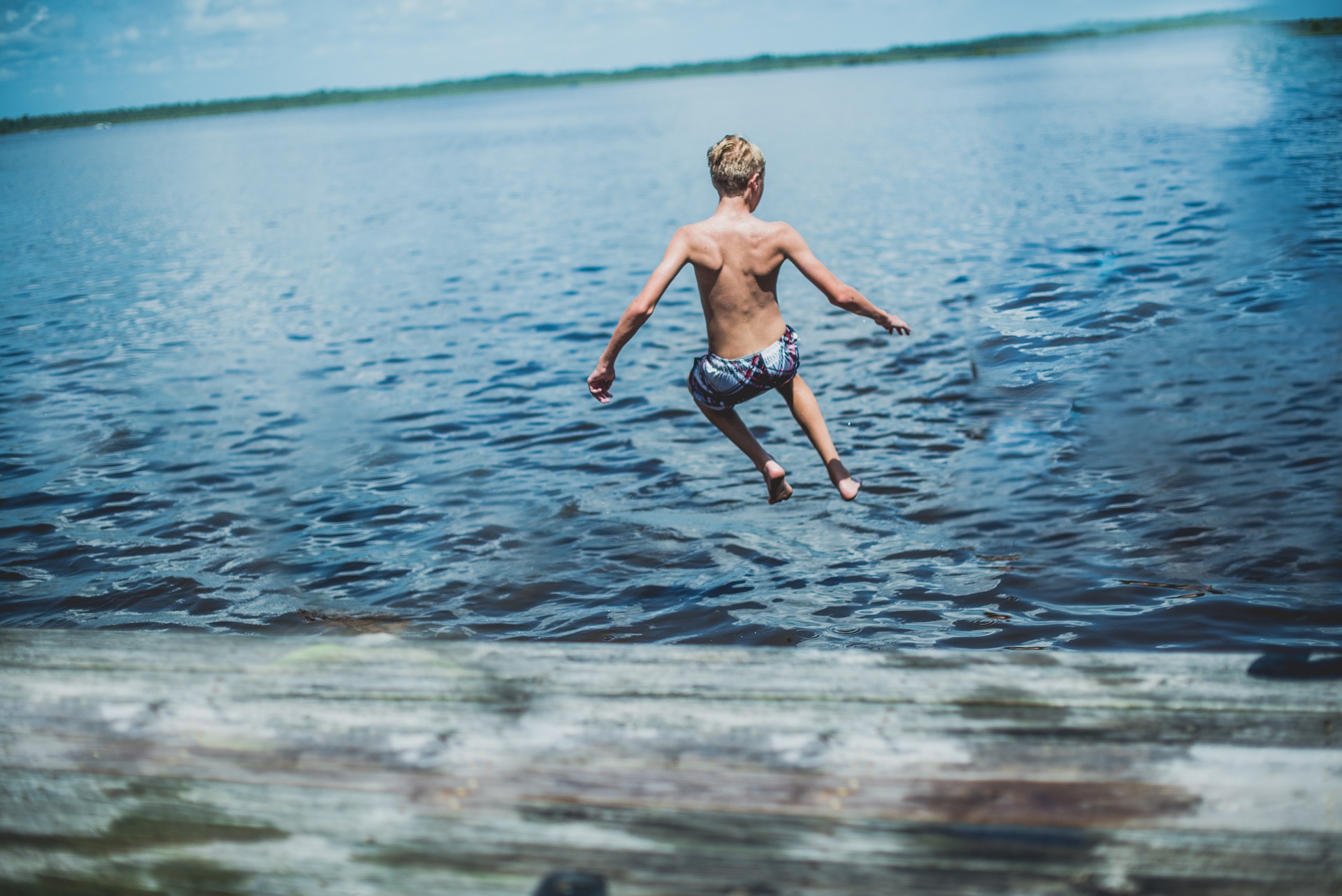 Ein Junge springt an einem Sommertag, von hinten gesehen, von einem Holzsteg in einen See.