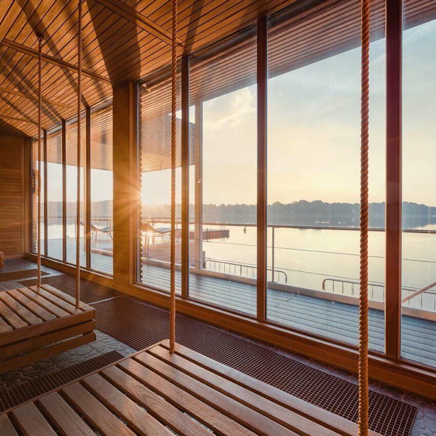 Hölzerne Schwebeliegen in einem Ruheraum der Fontane Therme mit Panoramablick auf den See bei Sonnenuntergang.