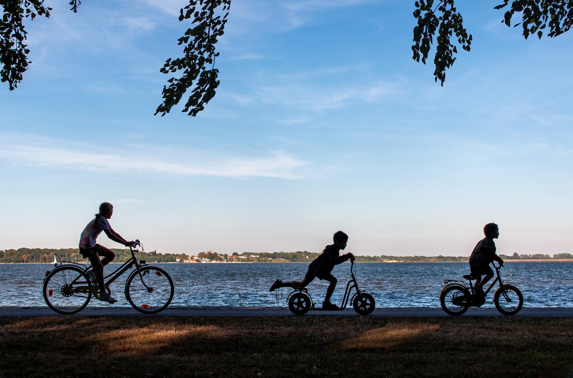Silhouetten von drei Kindern, die mit Fahrrad und Roller an einem Seeufer entlangfahren.