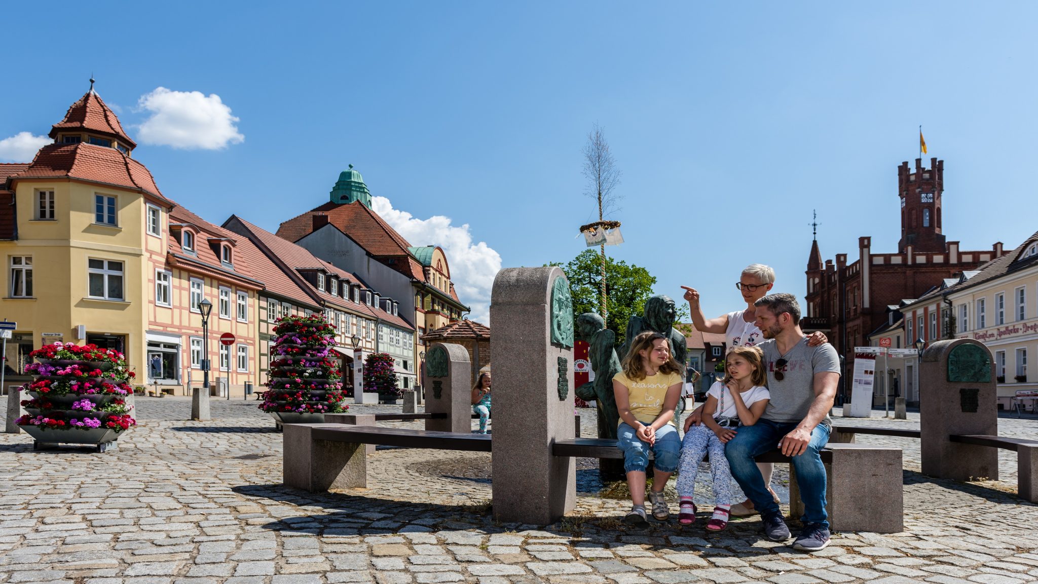 Eine Familie macht an einem sonnigen Tag eine Pause auf einer Bank auf dem historischen Marktplatz von Kyritz.