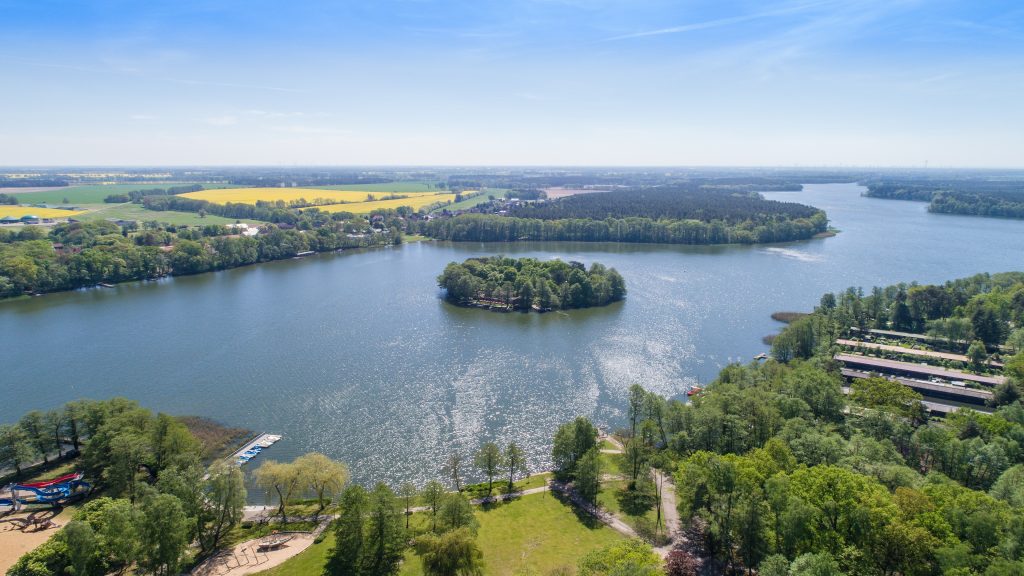 Luftaufnahme des Untersees bei Kyritz an einem sonnigen Tag mit der Insel in der Mitte und umliegenden Wäldern und Rapsfeldern.